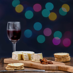 Brie and pecan halves on wooden cheese board, accompanied by crackers and one glass of red wine, against black background, decorated with colorful holiday lights- eating alone