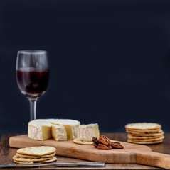 Brie and pecan halves on wooden cheese board, accompanied by crackers and one glass of red wine, against black background