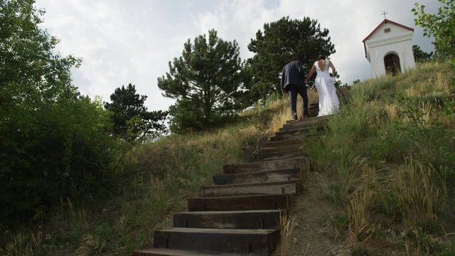 A newly married couple climbs a staircase to a shrine with a cross above.  4k. In slow motion.