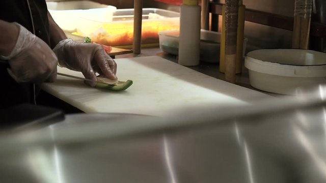 Process of making sushi rolls. Man cutting up green rite avocado by slices. Prepared sushi rolls pass through on foreground