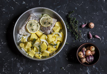 One pot lunch - white fish, potatoes, onions in a frying pan on a dark background. Raw ingredients for lunch. Top view