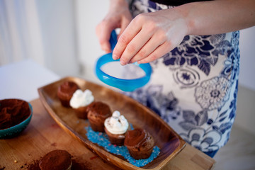 girl decorates cupcakes, holding plate, muffins and plate of ingredients for decoration on the table