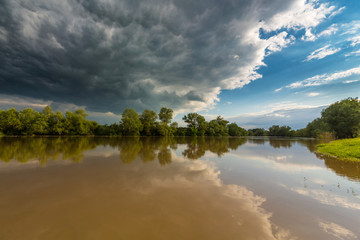 Ominous stormy sky over natural flooded river, with bright sun emerging from under the cumulus cloud cover