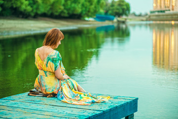 Red-headed woman in bright summer dress with open back sitting on the wooden pier at the river bank in the city. Reflection of buildings, trees and city lights in the water. Outdoor shot. Copy-space