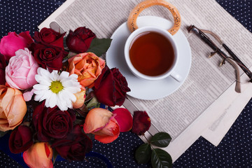 Beautiful vintage bouquet of roses with chamomile, newspaper and glasses. Cup of tea on saucer with bagels