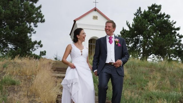 A newly married couple descends a staircase with a shrine and cross behind them. They are laughing. 4k. In slow motion.