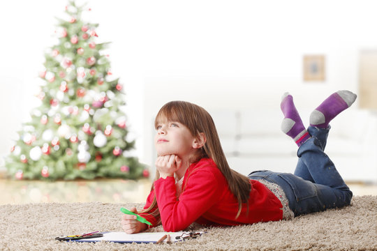 Lovely kid with christmas tree. Portrait of cute little girl lying on floor and coloring her book on christmas day..
