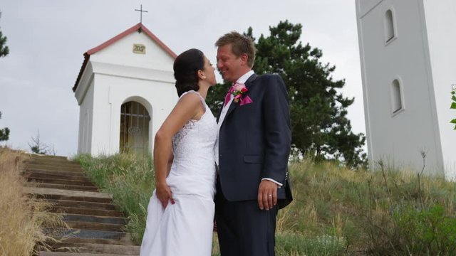 A newly married couple laugh as they look into each other's eyes on a staircase with a shrine and cross behind them. 4k. In slow motion.