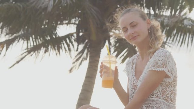 Young woman is sitting on the beach in sun and drinking cocktail through the straw.