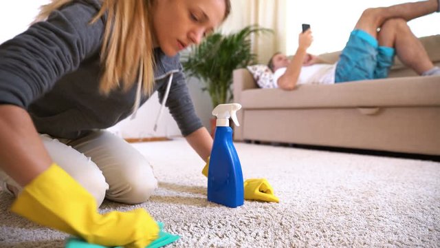 Tired wife cleans the carpet while her husband lies on the couch with the phone.