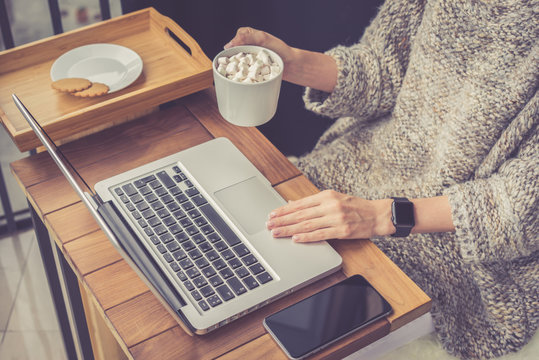 Girl in beige sweater looking for information on Internet using laptop while holding cup of cocoa with marshmallows.Woman sitting at wooden table on which is smartphone. Mock up. Life style.