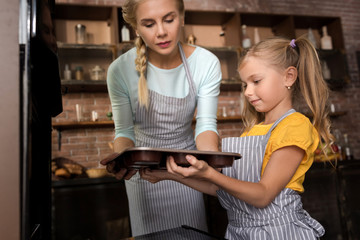 Involved girl putting with her mother pastry in the oven