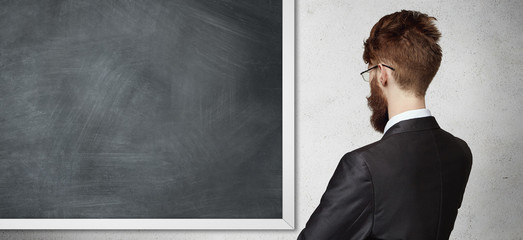 Back view of young employee wearing formal suit and glasses, standing in front of blackboard, presenting something. Bearded student or teacher staring at blank chalkboard during lesson in classroom