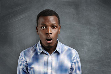 Headshot of puzzled or suprised African employee dressed in checkered shirt looking in shock and frustration at camera against blank chalkboard with copy space for your text or advertising content