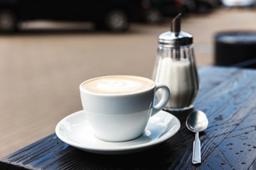 A white cup of cappuccino, sugar bowl and a tea spoon on the dark wooden table of a city cafe terrace
