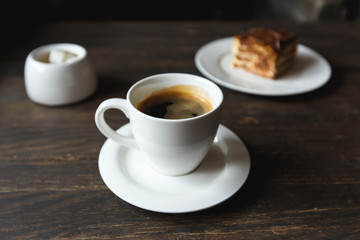 A white cup of black coffee, a piece of cake on the white plate, sugar bowl on the wooden table in a cafe. Selective focus, small depth of fieild.