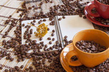 composition with open notebook and two cups filled with coffee beans standing on a newspaper and a wooden rug in shades of brown. Spilling coffee beans. Herbarium of yellow flower on a notebook page.