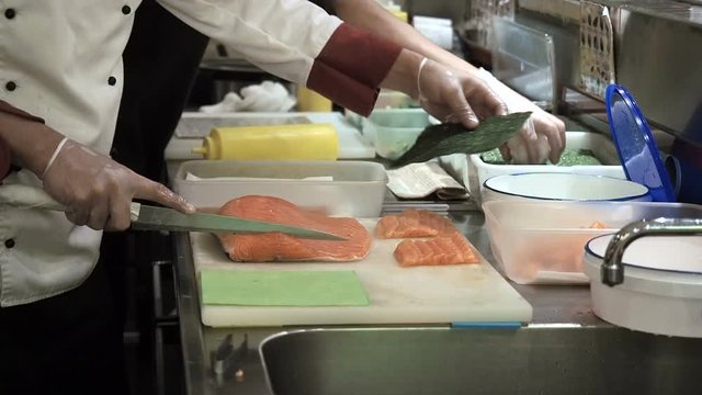 Process of making and cutting salmon for sushi rolls. Cook cutting up fish by knife on thin slices. Big red piece of trout meat laying on white cutting board. Japanese food and service