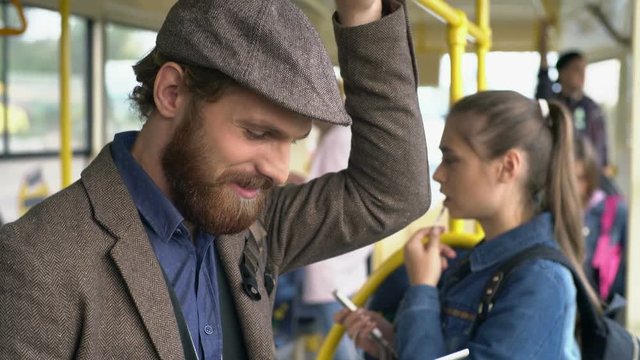 Tilt up of laughing young man with beard standing in trolleybus and communicating via tablet in his hand, young woman talking on phone in background