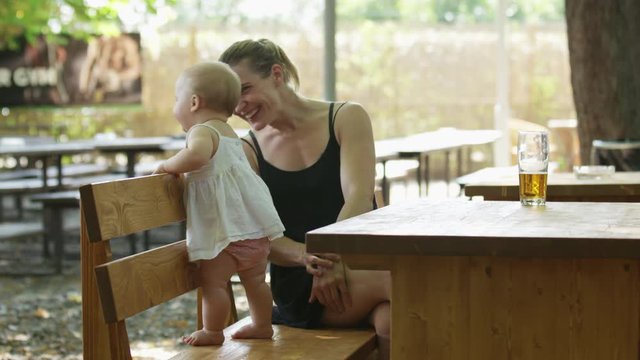 A mother sits in a beer garden with her baby standing on the bench beside her. The baby looks around while the mother smiles. 4k.