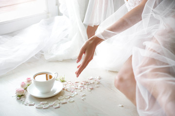 A picture of an elegant female hand with fine manicure pulling to a perfect cup of tea surrounded by rose buds, her bode covered with transparent cloth, blinding white light