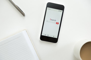 Top view of a mobile phone lying on a white office desk, notebook, mug with coffee, pen party seen. Business concept, lay flat