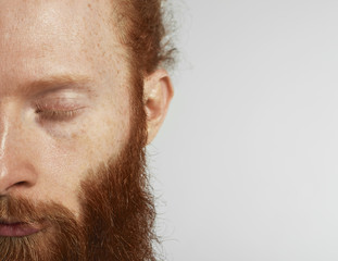 Close up half of face with closed eye. Red beard mans portrait in studio on grey background....