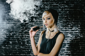 Sexy goth girl smokes electronic cigarette on dark background. The model vaper vaping a vaporizer...