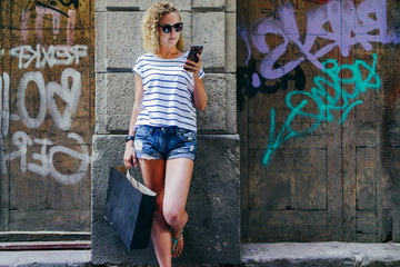 Obraz na płótnie Canvas Girl with blond curly hair, dressed in striped T-shirt and denim shorts,standing outside, leaning against brick wall and holding smartphone.Woman hand holds black bag with purchases.Girl is shopping.