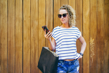 Girl with blond hair,dressed in striped T-shirt and denim shorts, standing with a black shopping bag while looking at screen of smartphone,is in her hand.In background wooden wall.Girl after shopping.