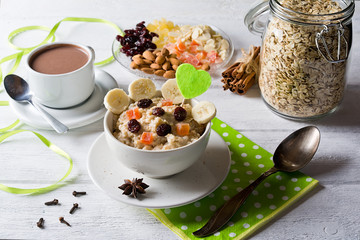 Oatmeal porridge with dried fruits, cranberries, bananas and spices. Cup of cacao and jar of oatmeal on background. Arranged on white background.
