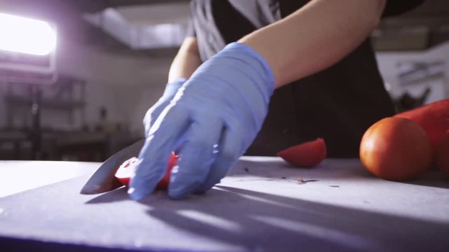 Hands of chef in gloves slicing up a tomato with a knife on cutting board