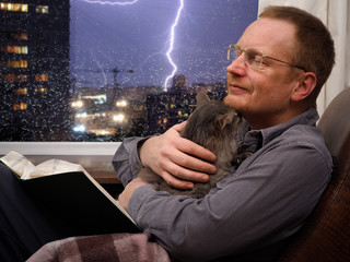 Man with a cat and a book in an armchair. Outside, night city, rain, storm