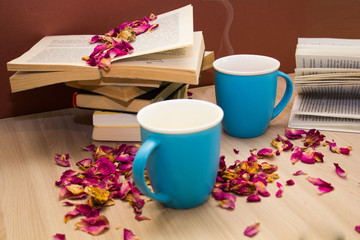 a stack of books and a hot drink in a blue mug on wooden table with rose petals