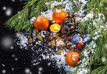 Fototapeta na wymiar Pine branches, candle, Christmas tree decorations, tangerines, nuts, the pine cone in snow on a black background 