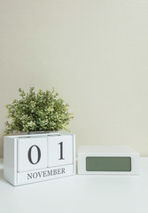 White wooden calendar with black 1 november word with clock and plant on white wood desk and cream wallpaper textured background , selective focus at the calendar