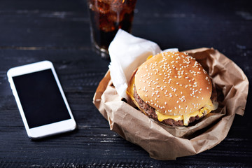 Cheeseburger in the paper container with paper napkins on a dark wooden background with moblie phone