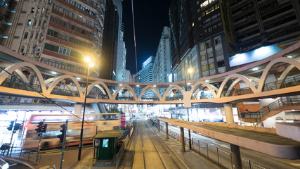 Night illuminated Hong Kong. View to tram stop, overground pedestrian bridge and moving double-decker bus among high-rise buildings