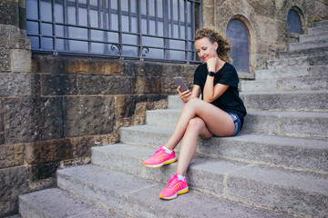 Summer day. Front view of smiling girl dressed in a black T-shirt, denim shorts and pink sneakers, sitting on the stone steps and surf the Internet on your phone. A young woman uses the gadget.