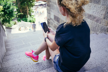 Summer day. Rear view, girl, dressed in a black T-shirt, denim shorts and pink sneakers, sitting on the stone steps and surf the Internet on your phone. A young woman uses the digital gadget.