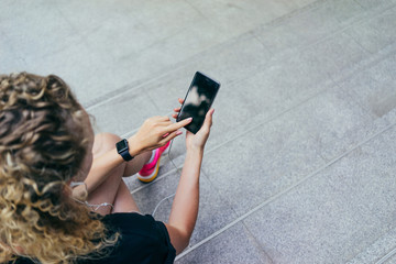 Summer day. Top view of a girl with blond curly hair sitting on stone steps and uses the smartphone with a blank screen. Young woman with headphones listening to music. Girl uses the gadget.
