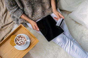 Girl dressed in knit sweater and white pants sitting in cozy atmosphere of home and choose Christmas gifts online store on digital tablet.Next is cup of cocoa with marshmallows. Mock up.Black screen.