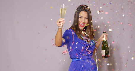 Sexy young woman celebrating New Year with a bottle and flute of chilled champagne as she dancing amidst falling confetti and colorful streamers