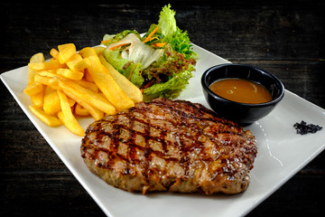 Plate with thick juicy stake, bowl of red sauce, golden french fries and green vegetables. Studio shot with black wooden background.
