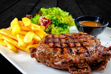 Plate with thick juicy stake, bowl of red sauce, golden french fries and green vegetables. Studio shot with black wooden background.