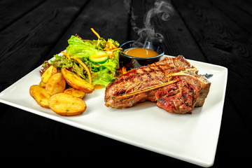 Plate with thick juicy stake, bowl of red sauce, golden french fries and green vegetables. Studio shot with black wooden background.