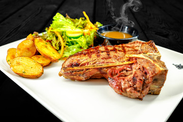 Plate with thick juicy stake, bowl of red sauce, golden french fries and green vegetables. Studio shot with black wooden background.