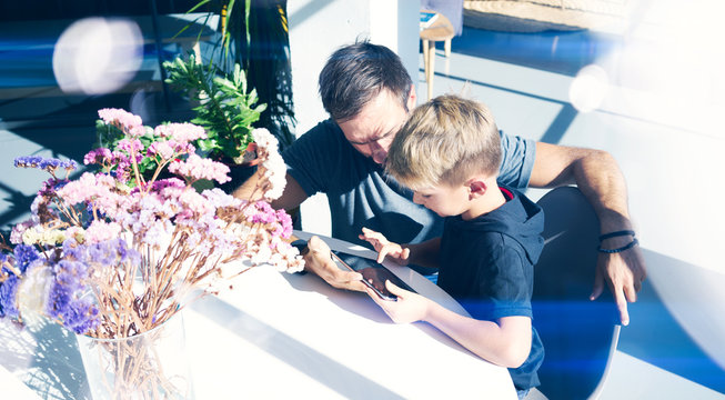 Young Father With His 7 Years Old Little Son Playing Digital Tablet At Sunny Home. Concept Of Happy Family Spending Time Together.Horizontal, Blurred Background.