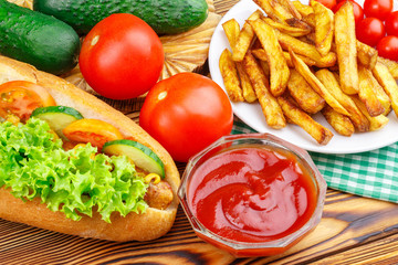 Fast food meal, hot dog, french fries, ketchup, tomato and cucumber on wooden background.