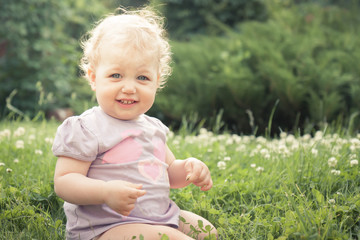 Smiling baby girl sitting in grass among blossoming flowers in summer park in sunny day with copy space